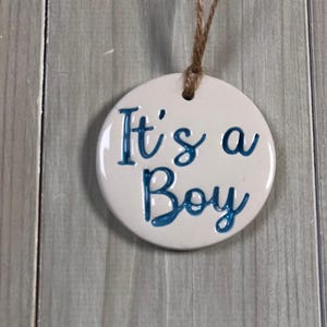May include: A circular, white ceramic ornament with the words "It's a Boy" in blue script. It is hung by a natural twine string, suitable for a baby shower or nursery decoration.
