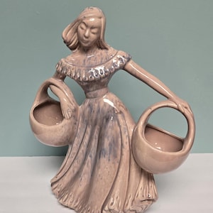 May include: A ceramic figurine of a woman in a long dress holding two baskets. The figurine is painted in shades of pink and purple.