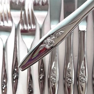 May include: A set of stainless steel flatware featuring a polished silver finish. Each piece is decorated with a raised, embossed rose motif on the handle. The collection includes multiple forks and knives arranged in a row, showcasing the floral detail.