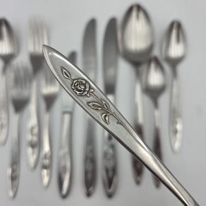 May include: Silverware set featuring a spoon with a rose and leaf design. The set includes forks, knives, and spoons, all with a similar floral motif. The silverware is arranged on a white surface, showcasing the elegant details.