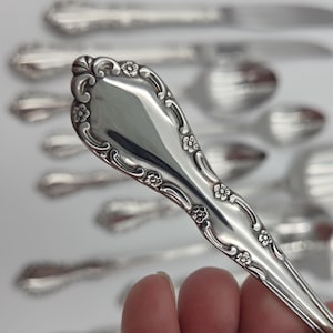 May include: A silver-toned metal spoon handle featuring an ornate embossed pattern with small floral motifs and scrolling edges. The handle has a polished, reflective surface and tapers toward the bowl, with additional blurred cutlery pieces visible.