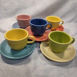May include: A collection of five ceramic tea cups and matching saucers in various solid colors including yellow, blue, pink, green, and orange. Each cup features a rounded body with horizontal ringed detailing and a curved handle on the side.