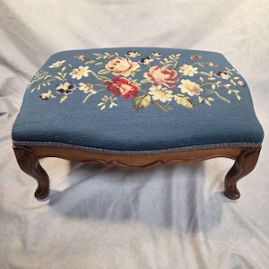 May include: A rectangular footstool featuring a blue needlepoint top with a floral pattern of red, pink, and white roses and small white flowers. The base is constructed from brown wood with four curved legs and a blue fabric trim border.