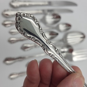 May include: A silver-toned metal spoon handle featuring an ornate embossed floral pattern with scrolling edges and a polished finish. The handle is tapered with a pointed end and decorative flower motifs along the sides, set against a blurred background.