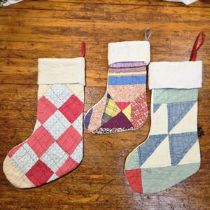 May include: Three quilted Christmas stockings with hanging loops. The left stocking features a red and light blue checkered pattern. The center stocking has a multi-colored patchwork design. The right stocking displays a blue and cream geometric pattern.