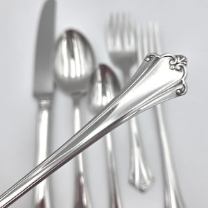 May include: A set of polished silver-toned flatware featuring a knife, two spoons, and two forks. The handle of the foreground piece displays a flared end with a decorative embossed scroll pattern, while the other utensils remain out of focus.