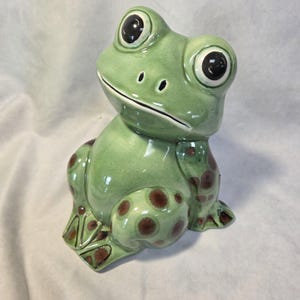 May include: A ceramic frog figurine with a glossy light green finish, featuring large black eyes and dark brown circular spots on its legs and feet. The frog is depicted in a seated position with a smooth, rounded body and a wide, curved mouth.
