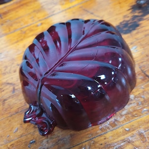 May include: A deep red, leaf-shaped glass dish. The dish has a detailed vein pattern and a glossy finish. The edges are gently curved, and the overall design is reminiscent of a natural leaf. The dish is sitting on a wooden surface.