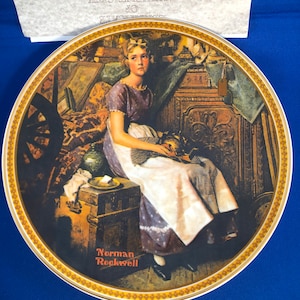 Puede incluir: Un plato de cerámica de estilo vintage con una pintura de una niña sentada en un baúl en un ático. La niña lleva un vestido azul y blanco y sostiene un pequeño animal de peluche. El plato tiene un borde dorado y la firma del artista, "Norman Rockwell", está impresa en rojo en la parte inferior.