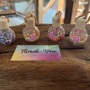 May include: Four small glass bottles with cork stoppers and colorful floral designs. The bottles are hanging from gold cords and are arranged on a wooden surface. A thank you card is in the foreground.