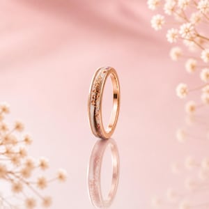 Op de afbeelding: Een roségouden ring met een witte band ingelegd met goudvlokken en kleine rode accenten. De ring staat tegen een zachtroze achtergrond met delicate witte bloemen, wat een romantische en elegante uitstraling creëert.