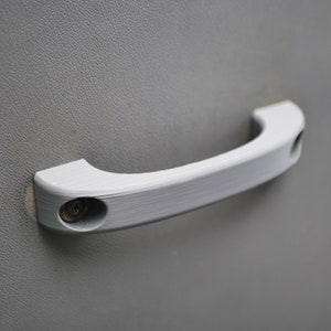 May include: Close-up of a light gray car door panel with two silver-colored handles. The main handle is a curved pull, and the other is a lever. The panel has a textured surface.