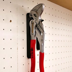 Puede incluir: Una llave ajustable de mango rojo con un soporte magnético negro montado en una pared de tablero de clavijas.