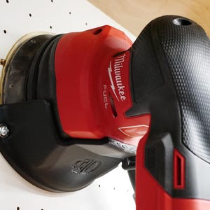 Puede incluir: Una lijadora orbital aleatoria Milwaukee FUEL roja y negra con una almohadilla de lijado de 12,7 cm. La lijadora está montada en un gancho de almacenamiento de pared negro.