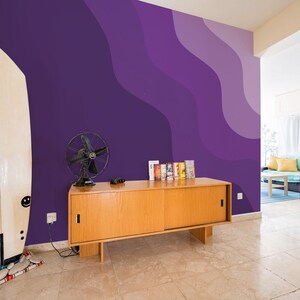 Könnte beinhalten: Ein Holzschränkchen mit zwei Türen steht vor einer violetten Wand mit wellenförmigen abstrakten Mustern. Ein weißes Surfbrett lehnt an der Wand links vom Schränkchen. Ein schwarzer Ventilator steht auf dem Schränkchen.