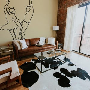 Peut inclure: Un intérieur de salon avec un canapé en cuir marron, un tapis en peau de vache noir et blanc, une table basse avec un plateau en verre et une table d'appoint avec une lampe. Le mur est peint en beige clair et présente un dessin au trait noir d'un couple dansant.