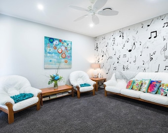 Op de afbeelding: Een woonkamer met witte muren en een wit plafond. Er is een witte bank met kussens en een witte fauteuil. De muur achter de bank heeft een muzieknotenbehang. Er hangt een schilderij aan de muur met blauwe, groene en rode kleuren.