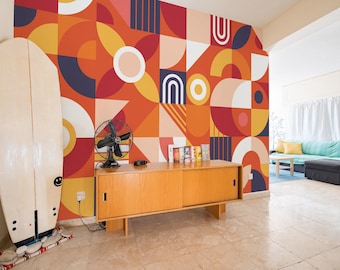 May include: A living room interior with a geometric patterned wall mural in shades of red, orange, yellow, and blue. A wooden cabinet with sliding doors is in front of the wall. A surfboard is leaning against the wall to the left of the cabinet. A fan is on the cabinet.