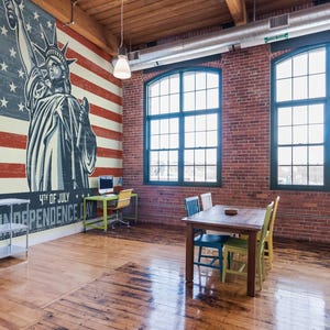 Puede incluir: Interior de un loft con paredes de ladrillo visto y grandes ventanales. Un mural de la bandera estadounidense y la Estatua de la Libertad domina una pared. La habitación está amueblada con una mesa, sillas y un escritorio.