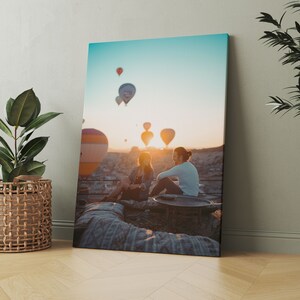 May include: A canvas print depicting a scenic sunrise with hot air balloons in the sky. Two people are seated, silhouetted against the sun. The print is propped against a wall, with a potted plant in a woven basket to the left.