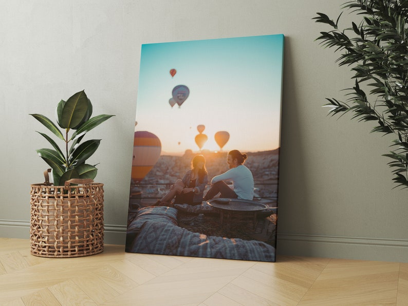 May include: A canvas print depicting a scenic sunrise with hot air balloons in the sky. Two people are seated, silhouetted against the sun. The print is propped against a wall, with a potted plant in a woven basket to the left.