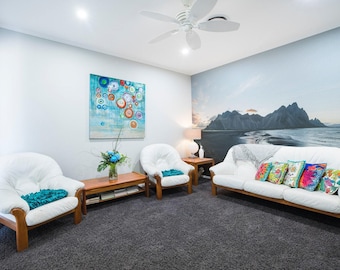 May include: A living room with two white armchairs and a white couch with colorful throw pillows. The wall behind the couch has a mural of a mountain range and a beach.