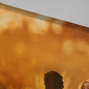 May include: Close-up of a canvas print corner, showcasing a warm, golden-orange color palette. The image features a blurred background with hints of figures, suggesting a landscape or portrait. The canvas is stretched and mounted on a wooden frame.