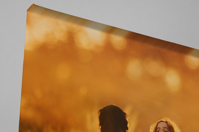 May include: Close-up of a canvas print corner, showcasing a warm, golden-orange color palette. The image features a blurred background with hints of figures, suggesting a landscape or portrait. The canvas is stretched and mounted on a wooden frame.