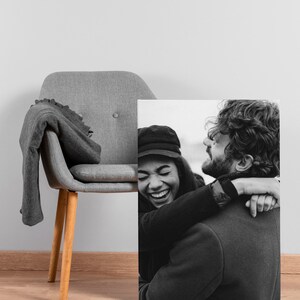 May include: Black and white canvas print of a joyful couple embracing. The woman is wearing a black hat and coat, laughing. The man has a beard and is wearing a dark coat. A gray chair with a blanket is in the background.