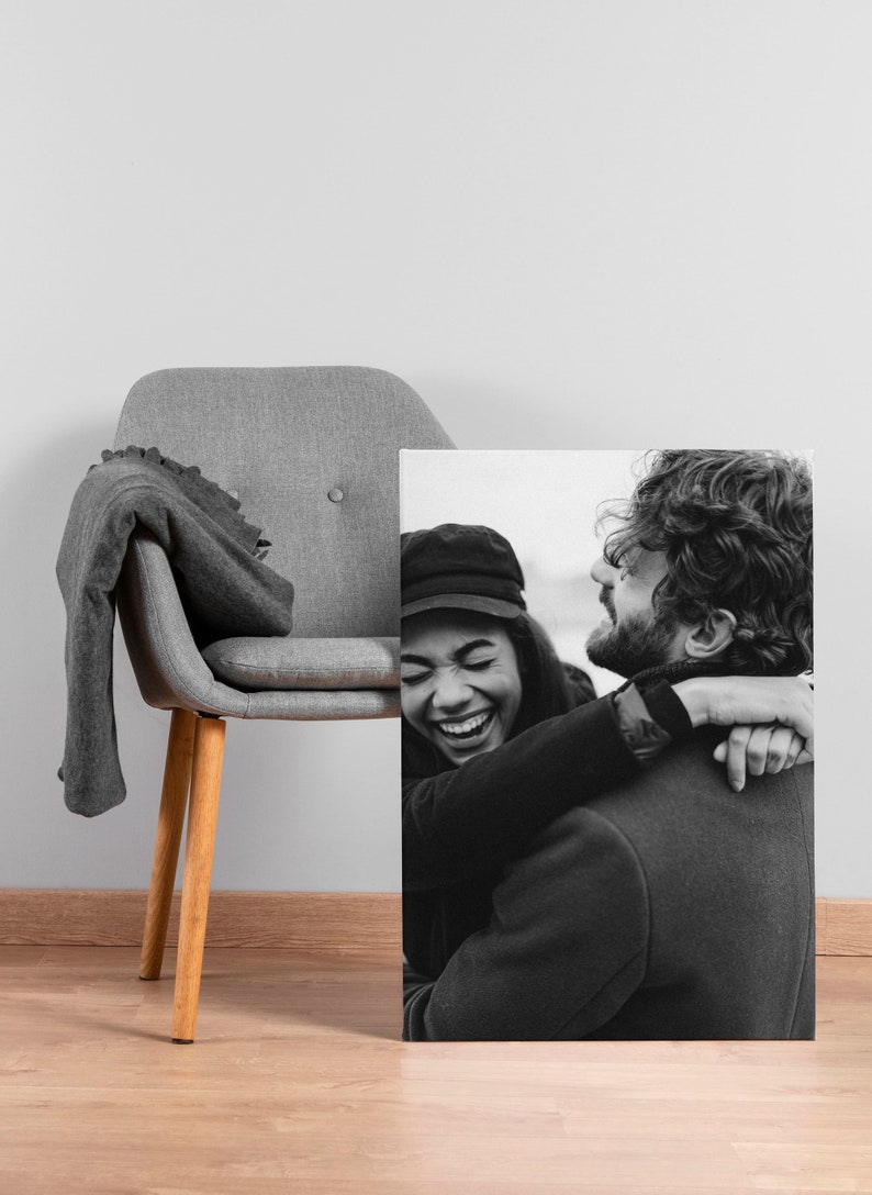 May include: Black and white canvas print of a joyful couple embracing. The woman is wearing a black hat and coat, laughing. The man has a beard and is wearing a dark coat. A gray chair with a blanket is in the background.