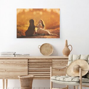May include: A canvas print featuring a girl with long hair, sitting with two dogs in a field. The dogs are a black dog and a golden retriever. The background is a warm, golden color, suggesting a sunset or sunrise.