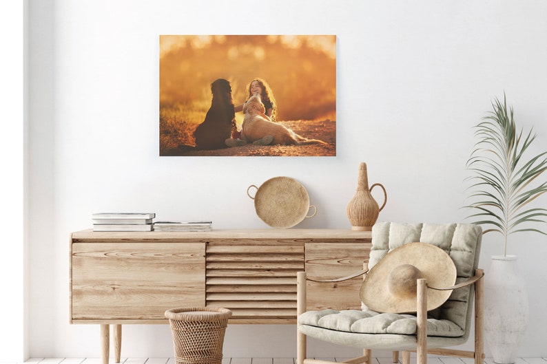May include: A canvas print featuring a girl with long hair, sitting with two dogs in a field. The dogs are a black dog and a golden retriever. The background is a warm, golden color, suggesting a sunset or sunrise.