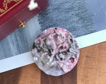 May include: A round, pink and gray gemstone pendant with carved floral and bird designs. The pendant has a hole at the top for a necklace. The pendant is displayed on a white surface with a red jewelry box and dried flowers.