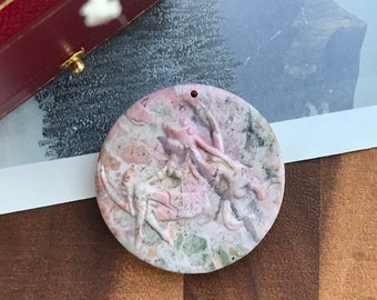 May include: A round, pink and green stone pendant with a carved dragon design. The pendant has a small hole at the top for a necklace. The pendant is on a wooden surface.