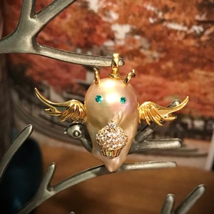 May include: A whimsical brooch featuring a pearl-like body with gold wings and a cupcake-shaped base. The brooch has teal-colored eyes and is suspended from a silver-colored branch. The background is a blurred view of fall foliage.