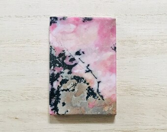 May include: A rectangular stone with a pink and black pattern. The stone has a polished surface and a natural, organic appearance. The pink color is dominant, with black and gray veining throughout. The stone is approximately 2 inches long and 1 inch wide.