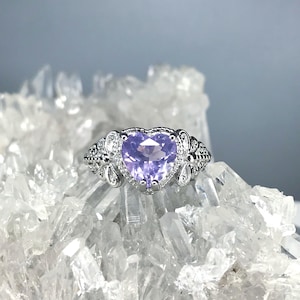 May include: A silver ring with a heart-shaped, light purple gemstone. The ring has intricate filigree detailing and small, clear accent stones around the heart. The ring is displayed on a bed of clear quartz crystals.