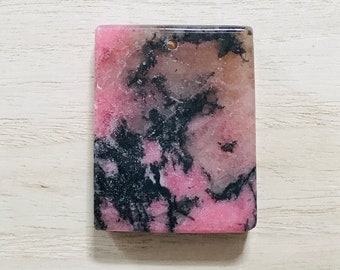 May include: A rectangular gemstone with a polished surface. The stone features a blend of pink, black, and brown hues, with a small hole drilled near the top. The colors are swirled and mottled throughout the stone.