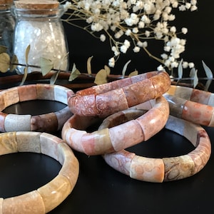 May include: Several coral fossil bracelets in shades of beige, brown, and pink are displayed. The bracelets are made of rectangular segments, creating a chunky, textured look. The background includes jars with cork lids and decorative sprigs.