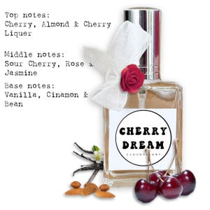 Peut inclure: Un flacon de parfum avec un bouchon argenté et une étiquette blanche indiquant "CHERRY DREAM". La bouteille est ornée d'un ruban blanc et d'une rose rouge. Des cerises fraîches, des amandes et des gousses de vanille sont disposées autour du flacon.