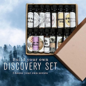 May include: A cardboard box containing multiple small glass vials with black caps, each featuring a unique label with text such as Here Dream, Blush Bloom, Whisper Spice, Gold H, and Oh So Sweet. The text Build your own DISCOVERY SET is visible.