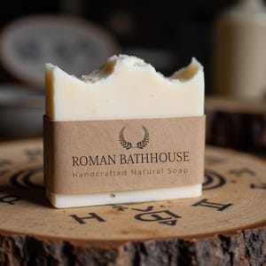 May include: A rectangular bar of handcrafted natural soap with a light beige color. The soap has a rough, uneven top edge. A brown paper band around the soap reads "ROMAN BATHHOUSE" and "Handcrafted Natural Soap". The soap rests on a wooden surface.