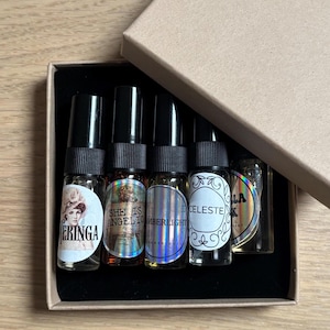 Pode incluir: Um conjunto de cinco pequenos frascos de vidro com spray e tampas pretas, dispostos dentro de uma caixa de papelão quadrada. Os frascos possuem etiquetas circulares com texto incluindo Eringa, She Is Angelic, Amberlight, Celeste e um adesivo holográfico.