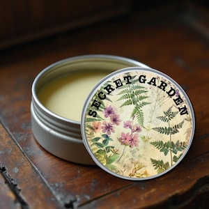 Op de afbeelding: Een kleine, open, zilverkleurige blikken doos met een lichtgele balsem. Het deksel toont de zwarte tekst "SECRET GARDEN" met een botanisch ontwerp van bloemen en varens. De doos staat op een donker houten oppervlak.