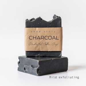 Op de afbeelding: Twee houtskoolkleurige zeepblokken, de ene bovenop de andere, met een gescheurde rand. Het bovenste blok is verpakt met een bruin papieren etiket met de tekst "soap story CHARCOAL Handcrafted Tallow Soap". De onderste balk heeft de woorden "Mild exfoliating" eronder.