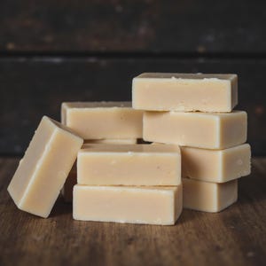 May include: A collection of rectangular, light-colored soap bars arranged on a wooden surface. The bars are stacked in a visually appealing arrangement, with some leaning against the stack. The background is a dark, textured surface.