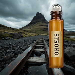 Könnte beinhalten: Eine braune Glas-Roll-on-Flasche mit silbernem Verschluss und einem Etikett mit der Aufschrift "STRONG" in Schwarz. Die Flasche steht vor dem Hintergrund eines Berges, eines steinigen Pfades und eines bewölkten Himmels.