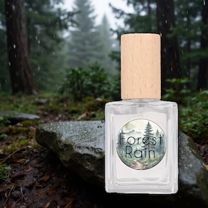 Könnte beinhalten: Eine klare Parfümflasche aus Glas mit hellem Holzverschluss. Das Etikett zeigt eine Waldszene mit den Worten "Forest Rain". Die Flasche steht vor dem Hintergrund eines üppigen, grünen Waldes.