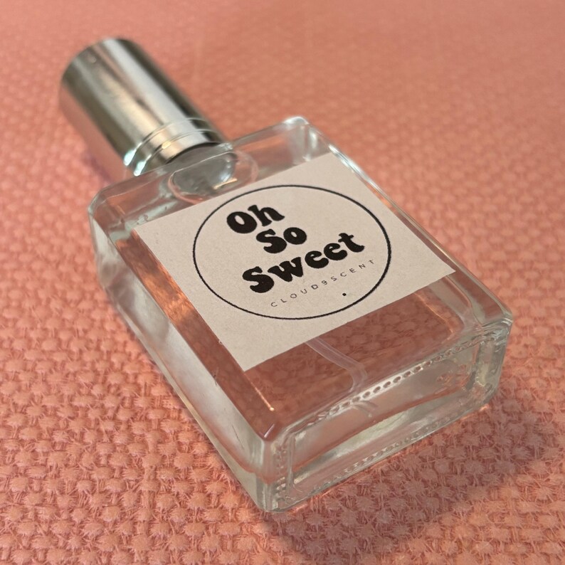 May include: A clear glass perfume bottle with a silver cap. The label reads "Oh So Sweet" in a circular design. The bottle contains a light pink liquid. The background is a textured pink surface.