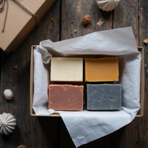 Könnte beinhalten: Geschenkbox mit vier rechteckigen handgemachten Seifen in den Farben Creme, Orange, Rotbraun und Grau. Die Seifen sind auf weißem Seidenpapier in einer braunen Pappschachtel angeordnet. Die Box ist mit braunem Bindfaden gebunden.